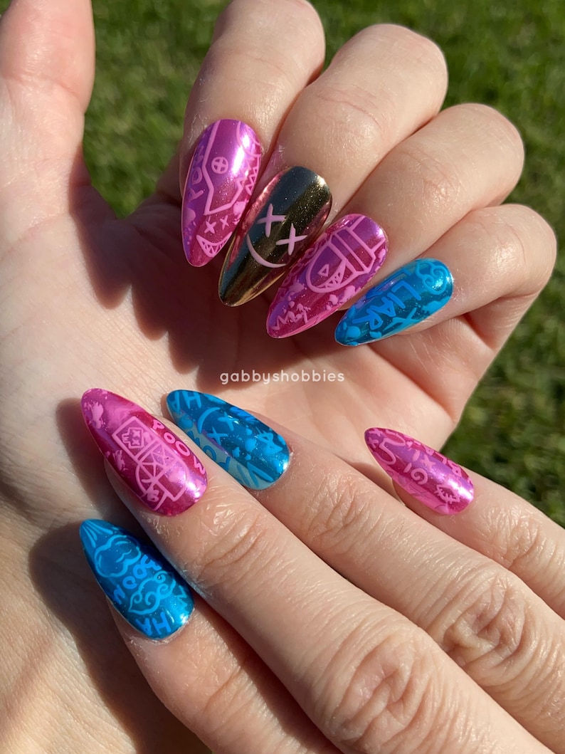 May include: A hand displaying a set of long, almond-shaped artificial nails. The nails feature a variety of designs, including pink and blue metallic finishes with intricate patterns and a gold nail with a black smiley face design.
