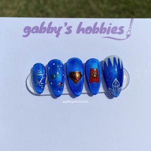 Pode incluir: Cinco unhas postiças azuis e brilhantes com vários desenhos. Uma unha apresenta um padrão geométrico prateado, outra tem um planeta e estrelas, uma terceira tem um emblema dourado, uma quarta tem uma personagem de desenho animado vermelha e a última tem um design de cristal branco e prateado. O texto "gabby's hobbies" é visível.