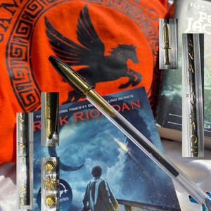 Pode incluir: Uma camiseta laranja Camp Half-Blood com um gráfico de pégaso preto e texto. Uma caneta transparente com detalhes dourados está sobre um livro de Rick Riordan. Itens adicionais incluem uma adaga dourada e capas de livros.