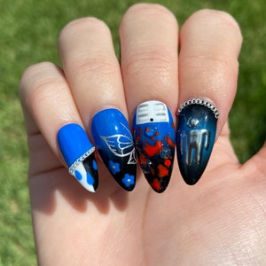 May include: Close-up of a hand with blue, black, and red nail art. The nails are painted with a variety of designs, including butterflies, flowers, and a small figure. The nails are also adorned with silver chain accents.