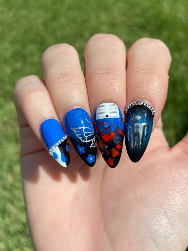 May include: Close-up of a hand with blue, black, and red nail art. The nails are painted with a variety of designs, including butterflies, flowers, and a small figure. The nails are also adorned with silver chain accents.