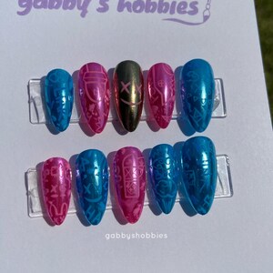 May include: A set of ten artificial nails in various colors, including blue, pink, and gold. The nails feature intricate designs and patterns. The text "gabby's hobbies" is visible at the top, and "gabbyshobbies" is at the bottom.