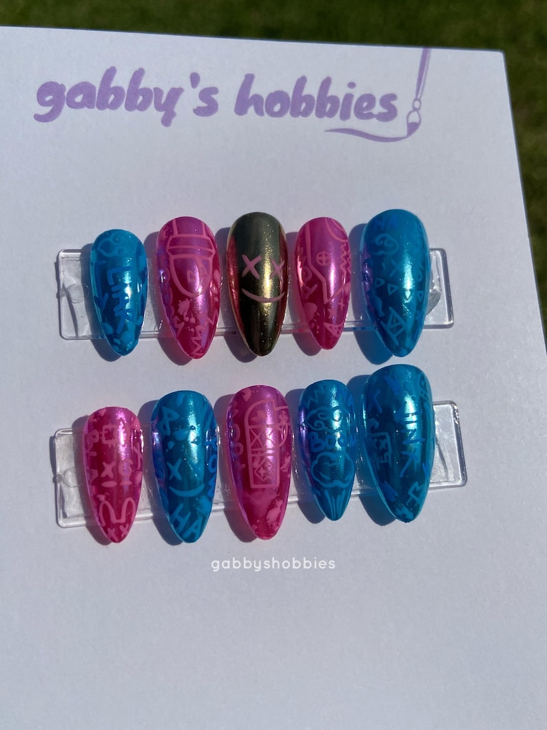 May include: A set of ten artificial nails in various colors, including blue, pink, and gold. The nails feature intricate designs and patterns. The text "gabby's hobbies" is visible at the top, and "gabbyshobbies" is at the bottom.