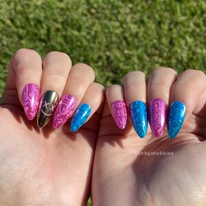May include: A close-up of two hands displaying long, almond-shaped artificial nails. The nails feature a variety of designs, including pink and blue patterns, a gold nail with a cross design, and a blue nail with a glossy finish. The background is blurred green grass.