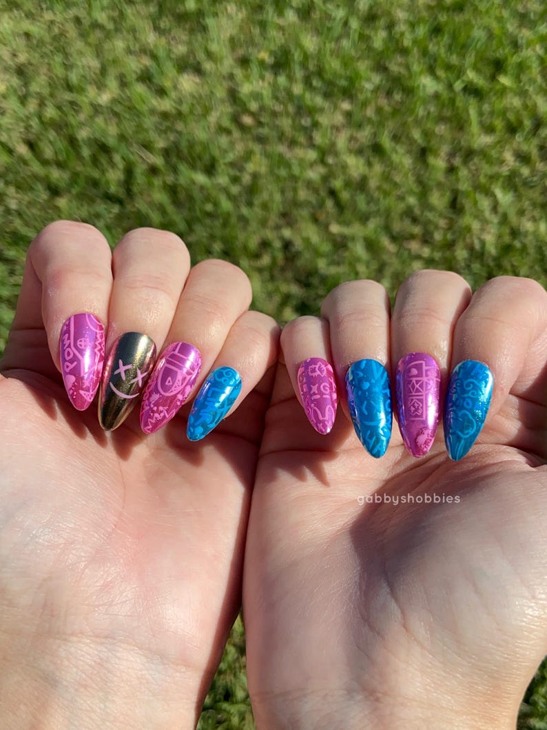 May include: A close-up of two hands displaying long, almond-shaped artificial nails. The nails feature a variety of designs, including pink and blue patterns, a gold nail with a cross design, and a blue nail with a glossy finish. The background is blurred green grass.