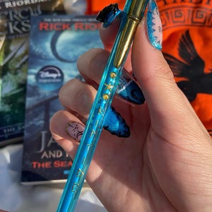 Op de afbeelding: Een blauwe en gouden pen met een decoratief ontwerp, vastgehouden in een hand met blauwe nail art. De pen heeft gouden accenten en kleine schelpenbedels. De pen heeft tekst op de schacht. Boeken zijn zichtbaar op de achtergrond.