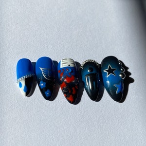 May include: Five blue and black nail art designs. The designs include a white teardrop with a silver accent, a blue and black floral design, a red and black abstract design, a black and silver design with a chain, and a blue and black star design with silver accents.