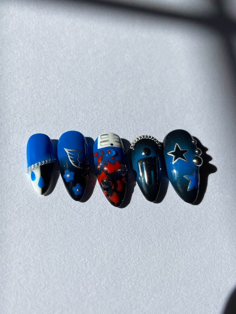 May include: Five blue and black nail art designs. The designs include a white teardrop with a silver accent, a blue and black floral design, a red and black abstract design, a black and silver design with a chain, and a blue and black star design with silver accents.