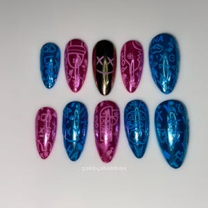 May include: A set of ten artificial nails in various colors, including blue, pink, and gold. Each nail features a unique design, with some displaying symbols and patterns. The nails are arranged on a white surface.