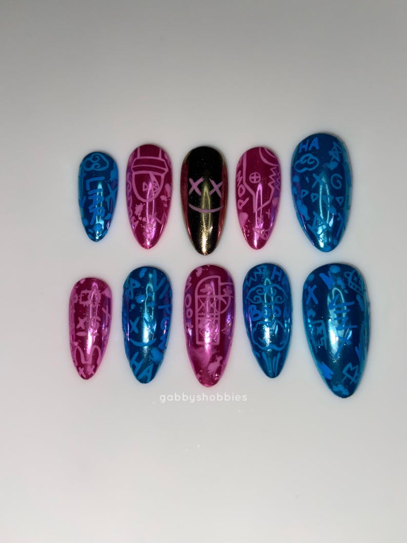 May include: A set of ten artificial nails in various colors, including blue, pink, and gold. Each nail features a unique design, with some displaying symbols and patterns. The nails are arranged on a white surface.