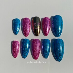 May include: A set of ten metallic press-on nails in various colors, including blue, pink, and gold. Each nail features a unique design, such as a mask or intricate patterns. The nails are arranged on a white surface. The text "gabbyshobbies" is visible.