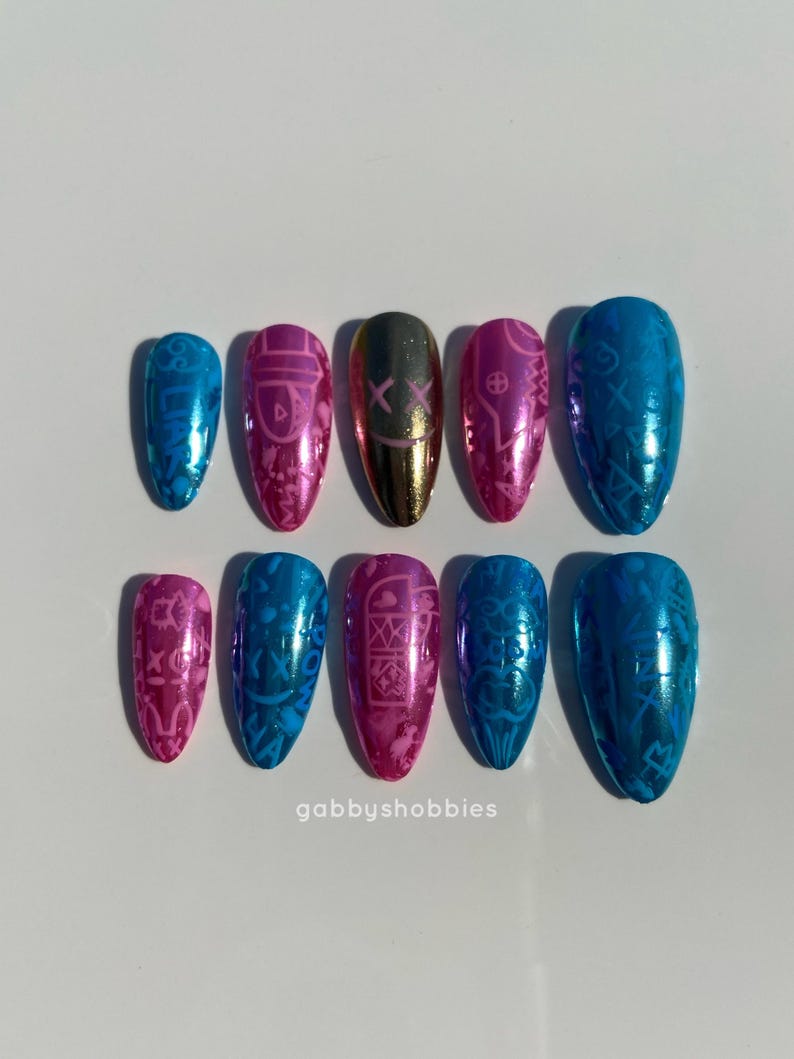 May include: A set of ten metallic press-on nails in various colors, including blue, pink, and gold. Each nail features a unique design, such as a mask or intricate patterns. The nails are arranged on a white surface. The text "gabbyshobbies" is visible.