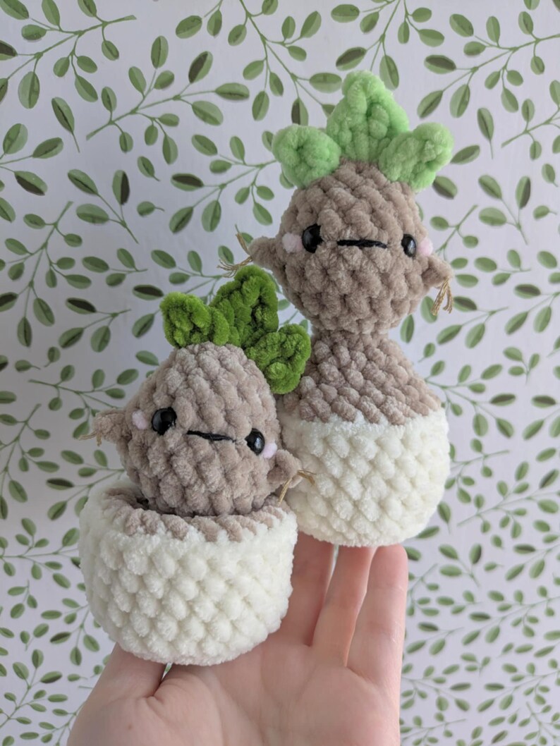 May include: Two crocheted plush toys shaped like potted plants. The plants are brown with green leaves and have cute faces. The pots are white.