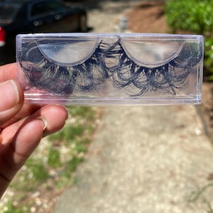May include: A pair of black false eyelashes in a clear plastic case. The eyelashes are long and full, with a wispy, layered design.