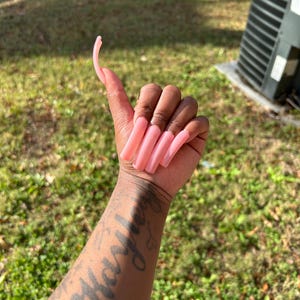 Könnte beinhalten: Eine Hand mit langen, hellrosa Acrylnägeln. Die Nägel haben eine einheitliche Farbe und reichen weit über die Fingerspitzen hinaus. Der Arm hat ein Tattoo mit dem Namen "Hayley" in Schreibschrift.