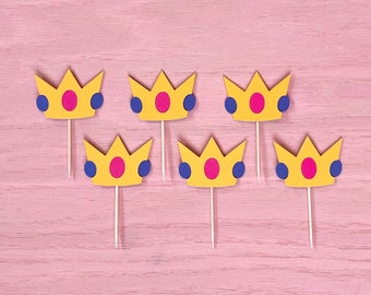 May include: Six yellow paper crowns with pink and blue circles on a pink wooden background. The crowns are attached to toothpicks.