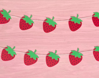 May include: A strawberry garland with ten red paper strawberries with green leaves strung on white string. The strawberries have small black seeds and are arranged against a pink wooden background.