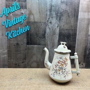 May include: Vintage white teapot with floral and butterfly designs. The teapot has a lid and a handle. The text "Aprils Vintage Kitchen" is in the upper left corner. The background is a wooden wall.