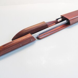 May include: A wooden sword scabbard with a partially exposed blade. The scabbard is made of reddish-brown wood and has a smooth, polished finish. The blade is silver-colored and appears to be made of metal. The scabbard is open, revealing the blade.
