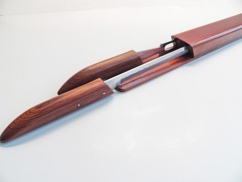 May include: A wooden sword scabbard with a partially exposed blade. The scabbard is made of reddish-brown wood and has a smooth, polished finish. The blade is silver-colored and appears to be made of metal. The scabbard is open, revealing the blade.