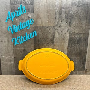 May include: A bright yellow Le Creuset oval baking dish. The dish has two handles and the number "32" is visible. The words "Le Creuset" are embossed on the bottom. The background includes the text "Aprils Vintage Kitchen".