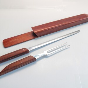 May include: A carving set with a long, slender knife and a two-pronged fork, both featuring wooden handles with silver rivets. A matching wooden case is also present. The set is designed for carving and serving meats.