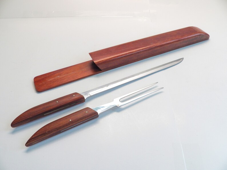May include: A carving set with a long, slender knife and a two-pronged fork, both featuring wooden handles with silver rivets. A matching wooden case is also present. The set is designed for carving and serving meats.