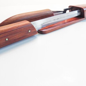 May include: A wooden sword scabbard with a partially exposed blade. The scabbard is made of reddish-brown wood with a visible grain pattern. The blade is silver-colored and appears sharp. Two small metal pins secure the scabbard.