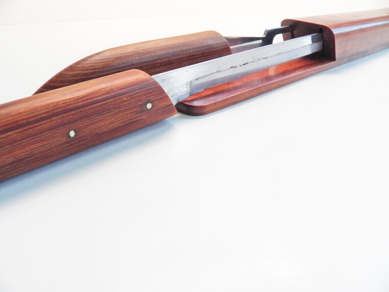 May include: A wooden sword scabbard with a partially exposed blade. The scabbard is made of reddish-brown wood with a visible grain pattern. The blade is silver-colored and appears sharp. Two small metal pins secure the scabbard.