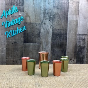 May include: A copper-colored pitcher and six small metal cups in shades of copper and green are arranged on a burlap surface. The words "Aprils Vintage Kitchen" are in turquoise script above the objects.