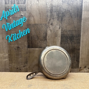 May include: A vintage, silver-toned metal frying pan with a long handle featuring three holes. The pan is resting on a burlap surface against a wooden background. The text "Aprils Vintage Kitchen" is in the upper left corner.