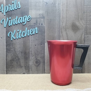 May include: A red vintage kitchen pitcher with a black handle. The pitcher is made of metal and has a glossy finish. The text "Aprils Vintage Kitchen" is displayed in light blue cursive letters.