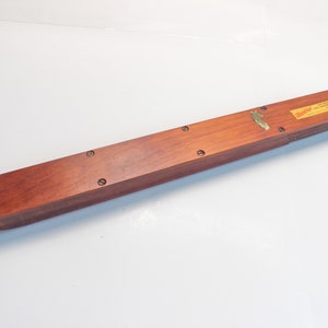 May include: A long, rectangular wooden box with a reddish-brown finish. The box has rounded ends and is secured with several screws. A small brass emblem and a rectangular label are affixed to the top. The label has text.