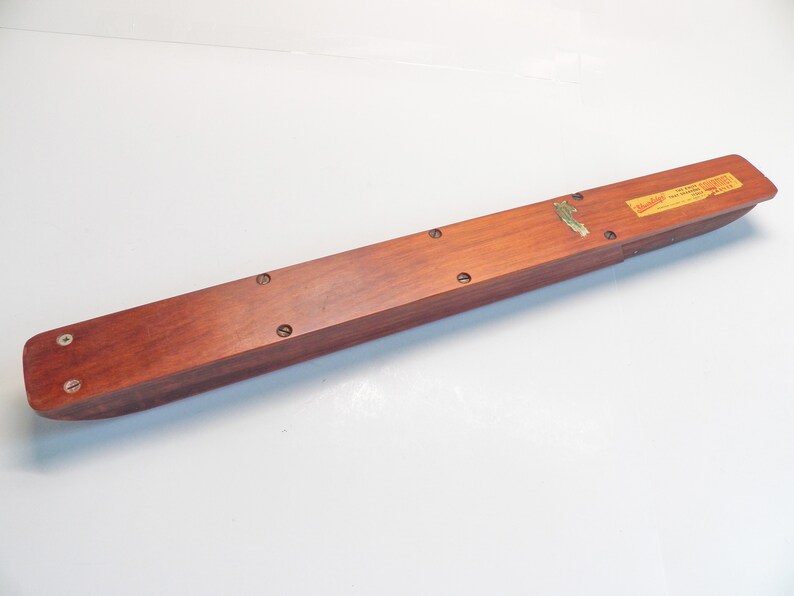 May include: A long, rectangular wooden box with a reddish-brown finish. The box has rounded ends and is secured with several screws. A small brass emblem and a rectangular label are affixed to the top. The label has text.