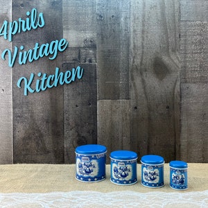 May include: A set of four blue and white ceramic kitchen canisters with lids. Each canister features a different label: Flour, Sugar, Coffee, and Tea. The canisters have a vintage design with a blue and white floral pattern and a scene of people.