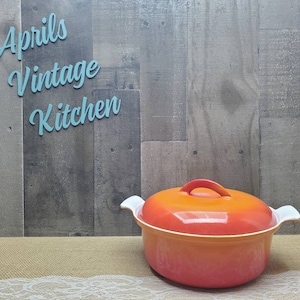 May include: An orange and red vintage cooking pot with a lid and white handles. The pot is on a burlap surface with the text "Aprils Vintage Kitchen" in the background.