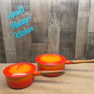 May include: Two orange and yellow enameled cast iron pots with lids and wooden handles. The pots are on a burlap surface. The text "Aprils Vintage Kitchen" is in the background.