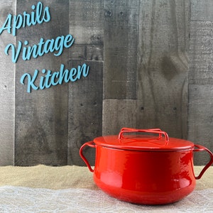 May include: A red enamel pot with a lid and handles, set against a gray wood-paneled backdrop. The words "Aprils Vintage Kitchen" are in light blue script above the pot. The pot is on a burlap and lace surface.