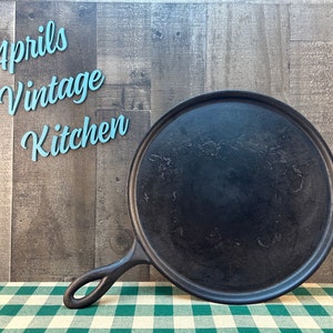 May include: A black cast iron griddle with a handle, perfect for cooking. The griddle is round and sits on a green and white checkered tablecloth. The words "Aprils Vintage Kitchen" are in the background.