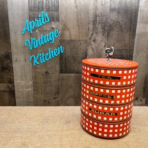May include: A cylindrical, stackable metal kitchen canister set featuring a red and white checkered pattern. The three tiers are labeled with black rectangular plates reading TEA, COFFEE, and SUGAR. A small metal rooster figure sits on the lid.