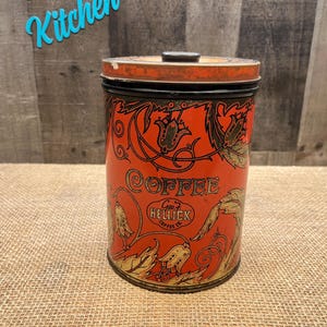 May include: Vintage red coffee tin with black and gold floral designs. The tin features the word "COFFEE" in gold lettering and the text "Geo. F. Heileck Coffee Co." This antique kitchenware item is cylindrical with a lid.