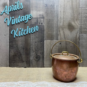 May include: A copper-colored vintage cooking pot with a lid and a brass handle. The pot is round with a slightly bulbous shape. The background is a wooden wall with the text "Aprils Vintage Kitchen" in light blue.
