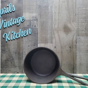 May include: A dark gray cast iron skillet with a long handle, resting on a green and white checkered tablecloth. The text "Aprils Vintage Kitchen" is in light blue script above the skillet. The skillet is a kitchenware item.