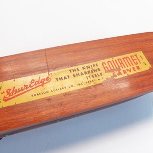 May include: A wooden knife sharpener with a rectangular shape and a reddish-brown finish. The label reads "ShurEdge Gourmet Carver" with the text "The knife that sharpens itself." The product is from RORESON CUTLERY CO., INC., PERRY, N.Y.