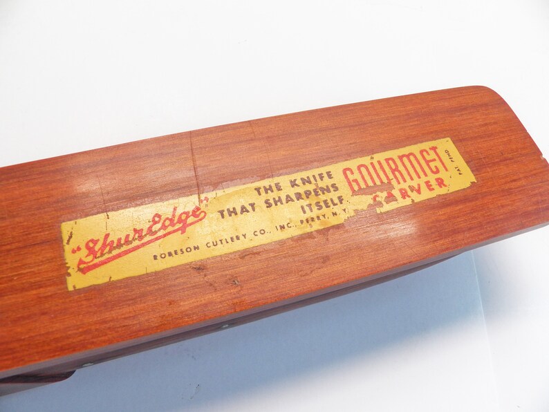 May include: A wooden knife sharpener with a rectangular shape and a reddish-brown finish. The label reads "ShurEdge Gourmet Carver" with the text "The knife that sharpens itself." The product is from RORESON CUTLERY CO., INC., PERRY, N.Y.