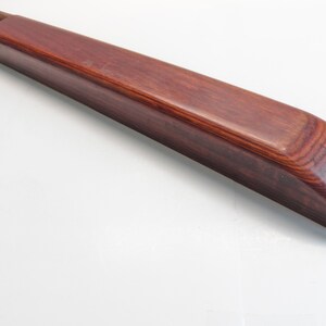 May include: A long, rectangular wooden object with a rich, reddish-brown finish. The item has a smooth, polished surface and rounded edges. Two cylindrical pieces protrude from one end. The wood grain is visible, adding to the natural aesthetic.