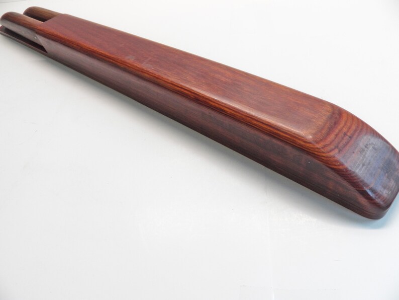 May include: A long, rectangular wooden object with a rich, reddish-brown finish. The item has a smooth, polished surface and rounded edges. Two cylindrical pieces protrude from one end. The wood grain is visible, adding to the natural aesthetic.