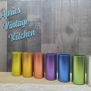 May include: A collection of seven metallic cylindrical containers in a rainbow of colors: yellow, orange, copper, purple, blue, green, and silver. The text "Aprils Vintage Kitchen" is displayed above the containers.