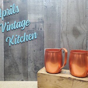 May include: Two copper-colored mugs with handles sit on a wooden box. The mugs have a shiny finish and a rounded shape. The words "Aprils Vintage Kitchen" are in light blue script above the mugs.