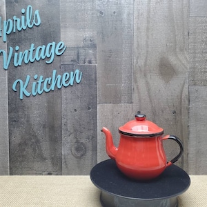 May include: A red enamel teapot with a black handle and trim, sitting on a black stand. The teapot has a lid with a black knob. The words "Aprils Vintage Kitchen" are in light blue script in the background.